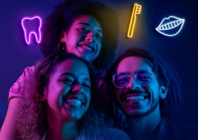 Três jovens sorrindo sob luzes de néon, com ícones luminosos de dente, escova e lábios, representando cuidados odontológicos da campanha Julho Neon da Hapvida +odonto.
