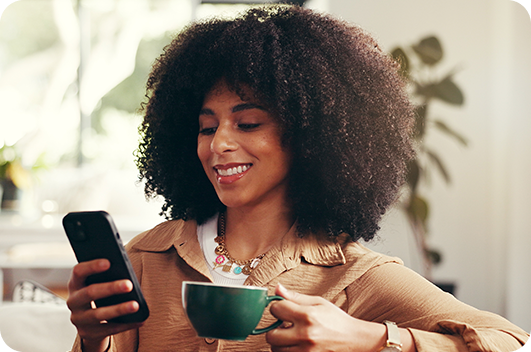 Mulher sorri enquanto usa o celular e segura uma xícara de café.